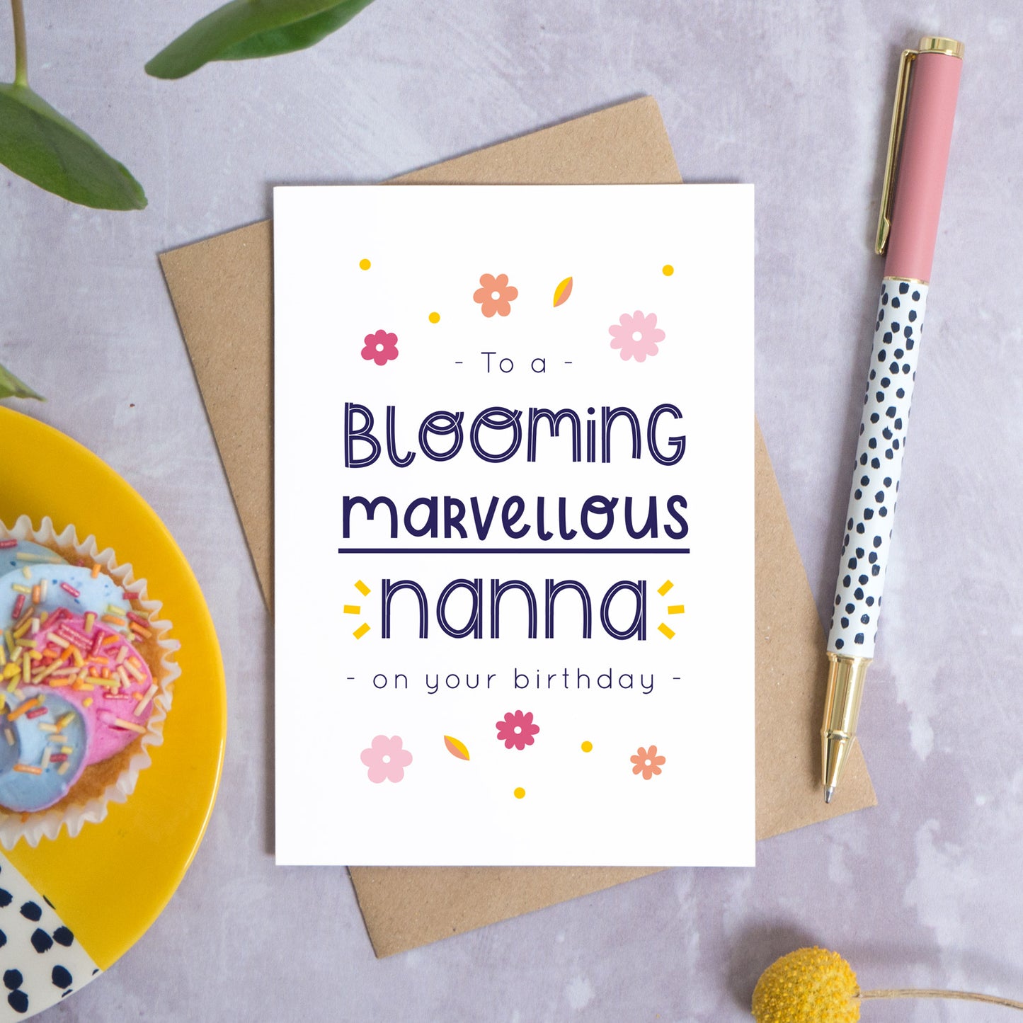 A blooming marvellous nanna birthday card photographed on top of it’s kraft brown envelope set on a grey background. To the left is a yellow plate with a birthday cupcake and to the top is green foliage. To the right is a spotty pen for scale and a pop of a yellow flower in the bottom right.