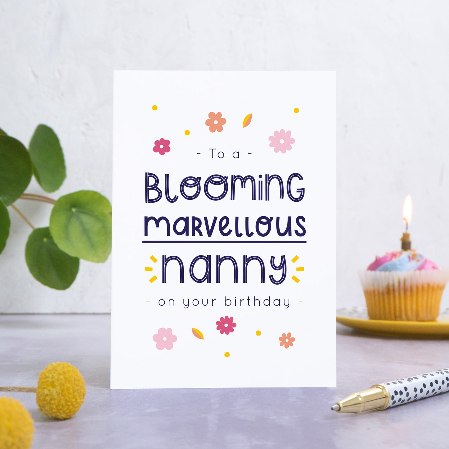 A blooming marvellous nanny birthday card photographed standing in front of a white background. In the background is a plant and a birthday cupcake and with a candle. In the foreground are two yellow flowers and a spotty pen.