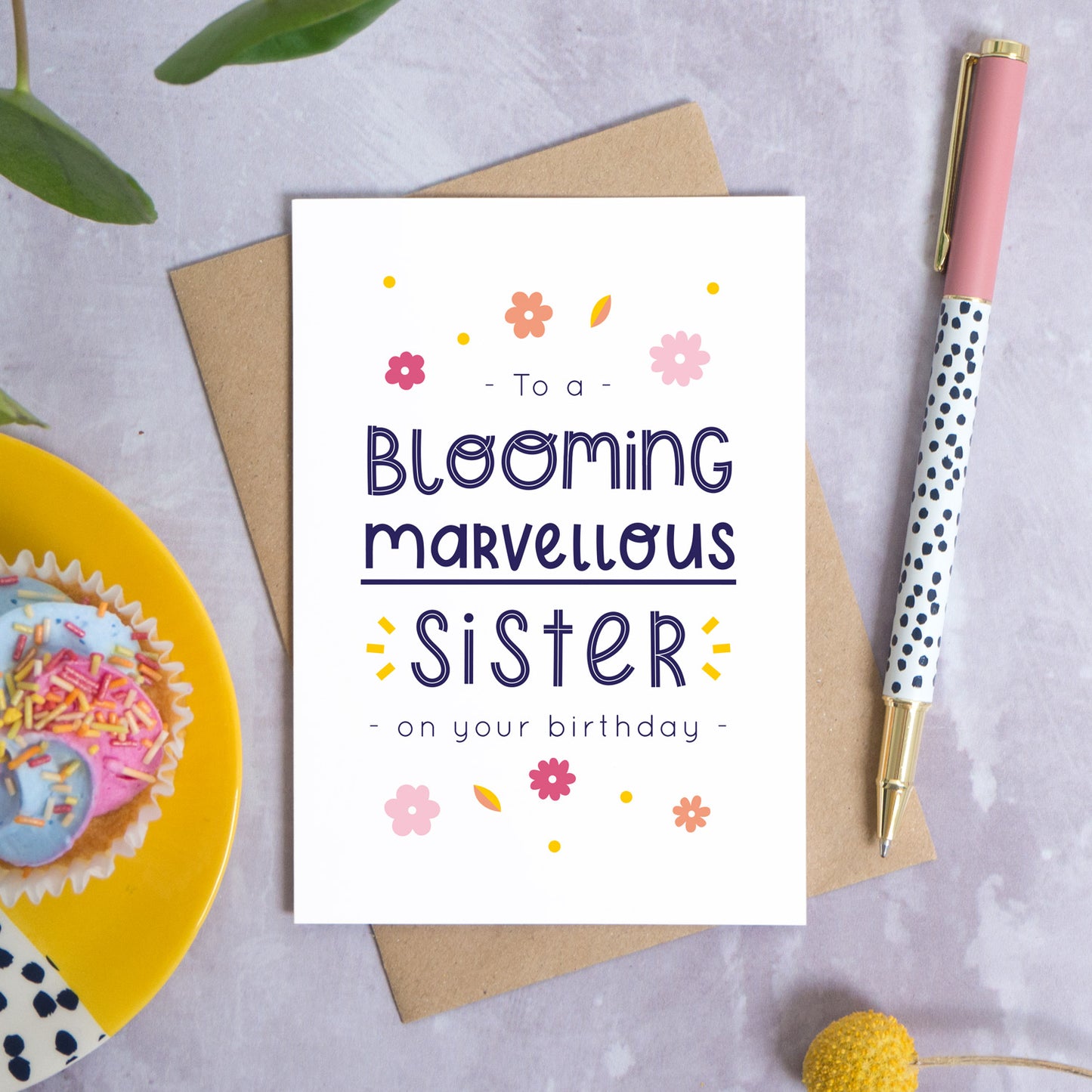 A blooming marvellous sister birthday card photographed on top of it’s kraft brown envelope set on a grey background. To the left is a yellow plate with a birthday cupcake and to the top is green foliage. To the right is a spotty pen for scale and a pop of a yellow flower in the bottom right.