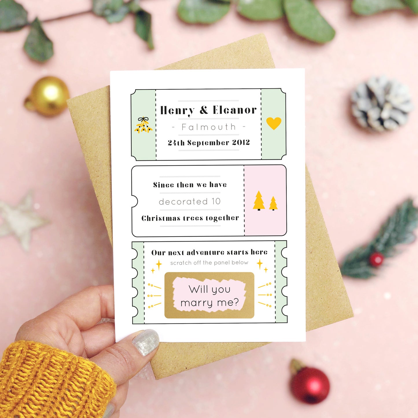 A personalised will you marry me christmas scratch card in pink and green held over a pink background with green foliage and gold and red baubles. The card is split up into tickets and the scratch panel has been scratched off to reveal the words ‘will you marry me?’.
