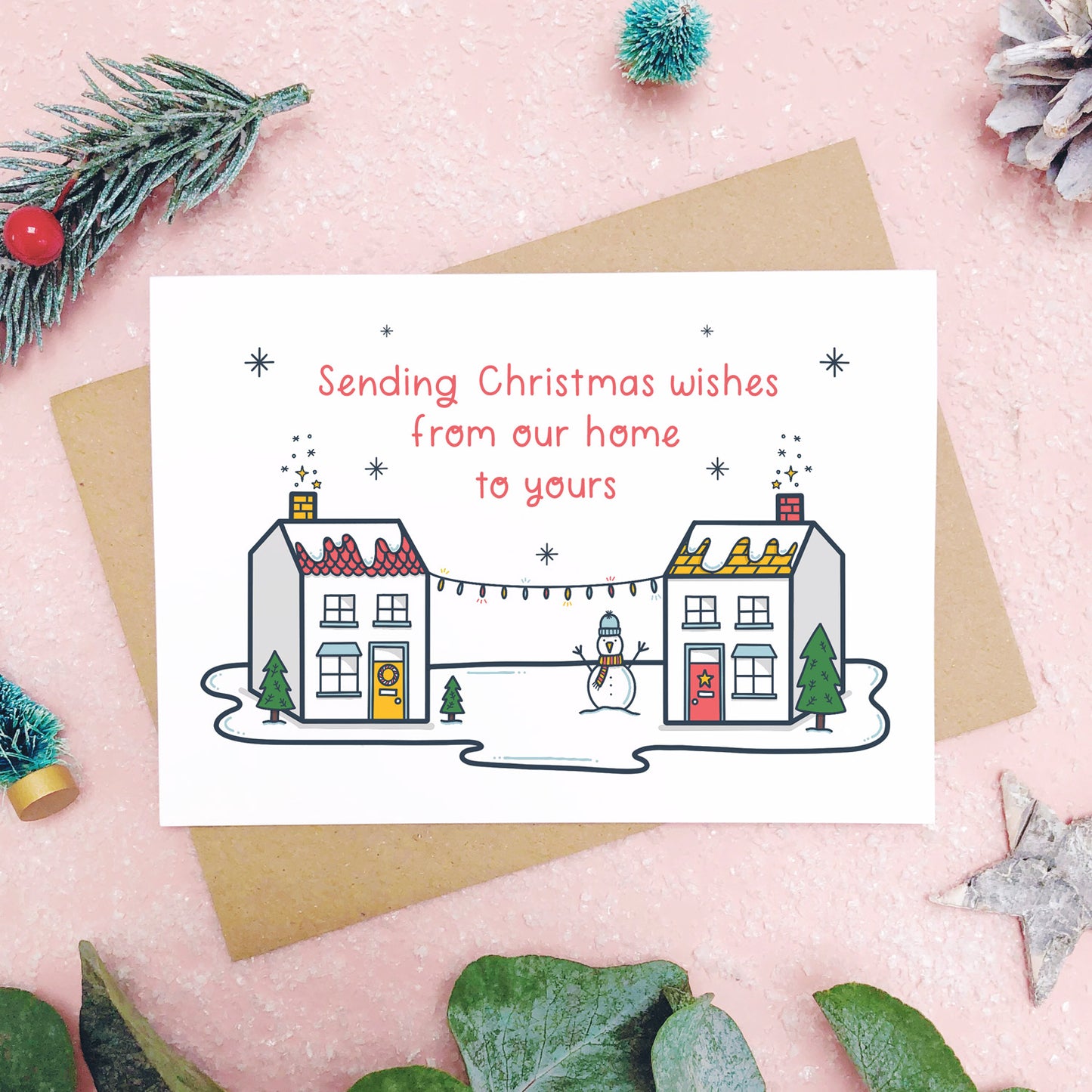 A sending wishes from our home to yours card photographed on a pink background with grey and green foliage. The card features two little houses connected by fairy lights.