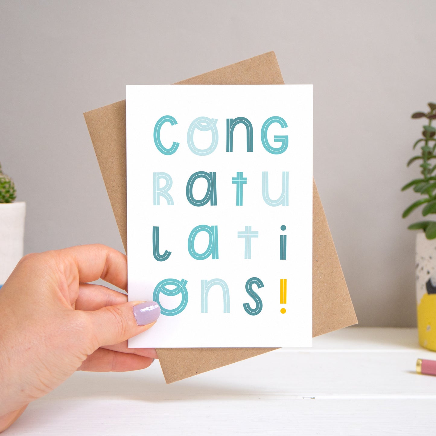 A congratulations card held over a warm grey and white background with potted plants peeping the sides. Behind the card is a kraft brown envelope that comes with the card. The text on this version of the card is in varying tones of blue.