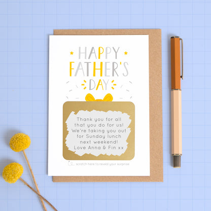 A personalised Father’s day scratch card photographed on a blue background with a pop of yellow flowers and a pen for scale. This image shows the grey and yellow version of the card with the golden present scratched off to reveal the message.