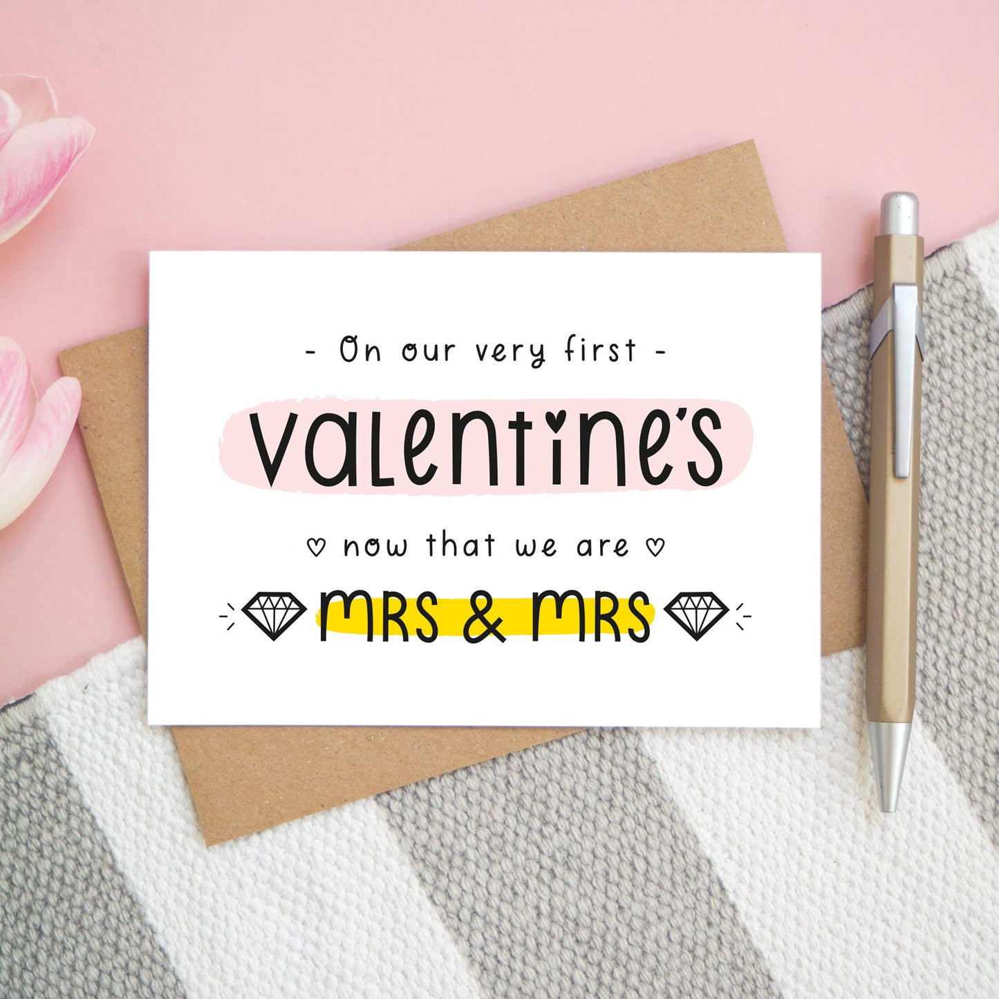 A first anniversary or Valentine’s card photographed on a pink background with pink tulip flowers, a gold pen and a grey and white stripe rug. This image shows the first valentine’s option with the Mrs & Mrs wording. The text is black and there are pops of yellow and pink behind key words.