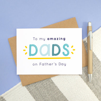 An amazing dads card laying on a stripy carpet and a blue background. The card shows navy and varying tones of blue text with a yellow underline and flicks. The card reads: "to my amazing dads on Father's day"