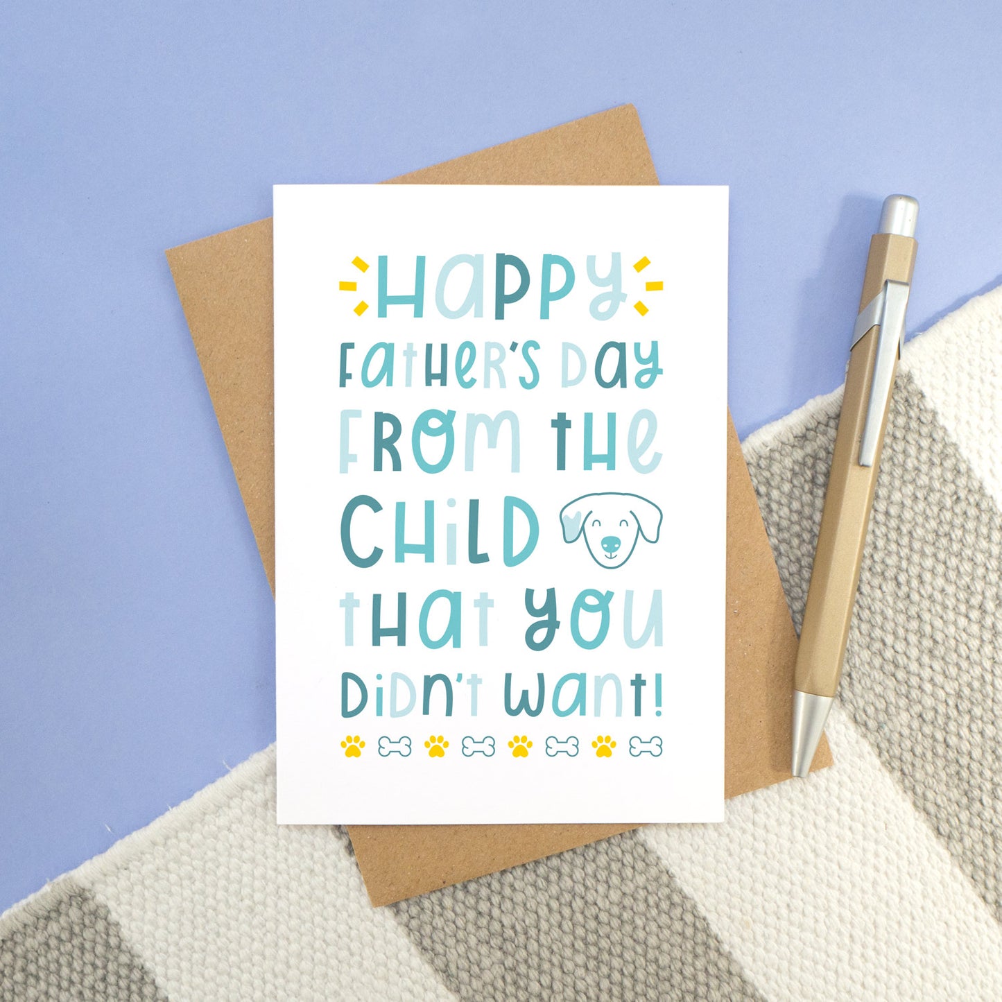 A dog child you didn't want card laying on a stripy carpet and a blue background. The card has varying tones of blue text, a dog illustration, paw prints and bones.