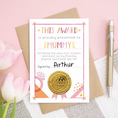 A Mummy certificate card for mother's day featuring a shiny gold seal. This card is pink and peach in colour with a small pop of yellow and has been shot over head on a pink and grey and white background. There is a gold pen for scale and tulips on the left.