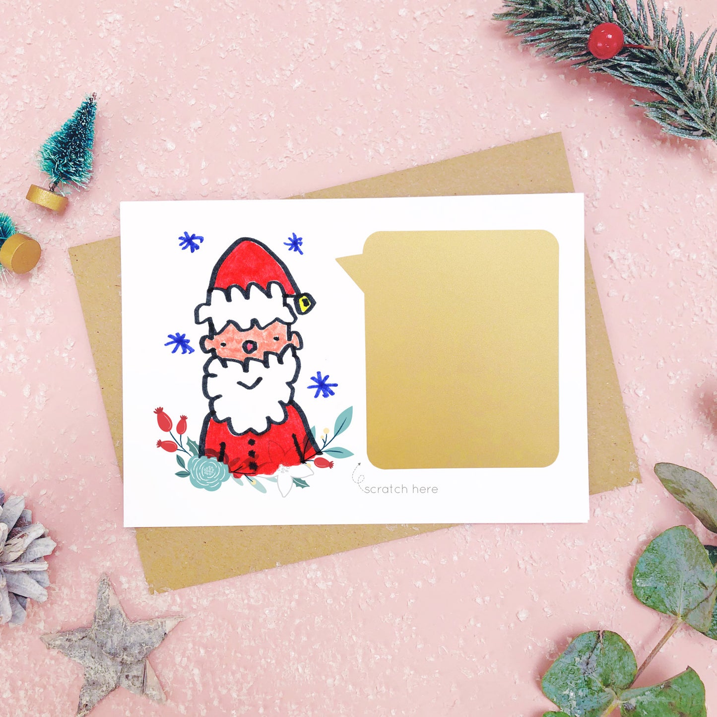 A make your own scratch card with a childs drawing of santa and an example of the panel after it has been applied. Shot on pink with snow and greenery.