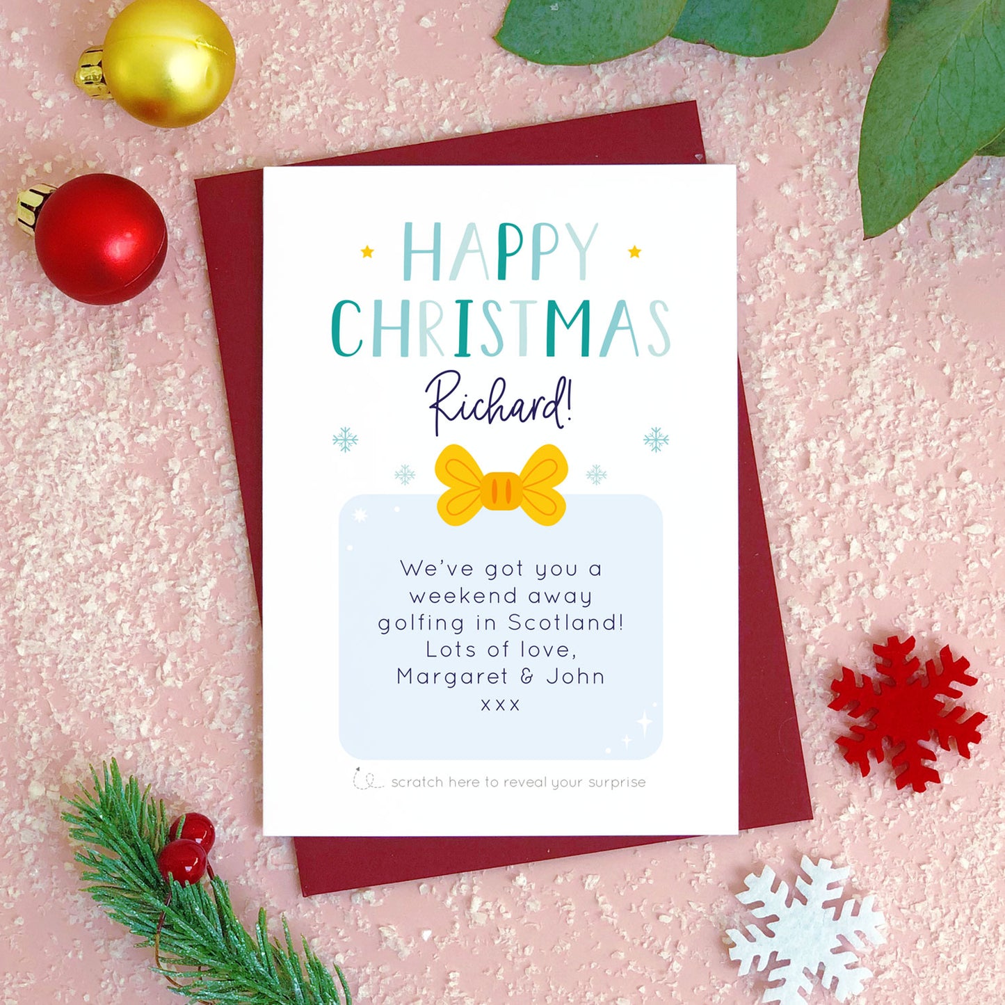 A personalised present christmas scratch card photographed flat lying on a red wine coloured envelope on a pink surface surrounded by fake snow, baubles and foliage. The gold panel has been completely scratched away to reveal a personalised message and gift reveal. This is the blue version of the card.