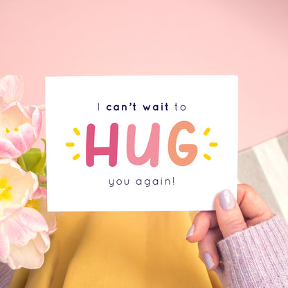 The non personalised, 'I can't wait to hug you again' card in pink. It has been shot on a pink background and has tulips to the left of the photo and is being held in the right.