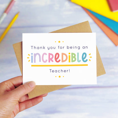 An incredible teacher thank you card being held over a blue background with colourful text books and two pens. The card is sat on top of its kraft brown envelope. This is the teacher card with the rainbow text colour option.