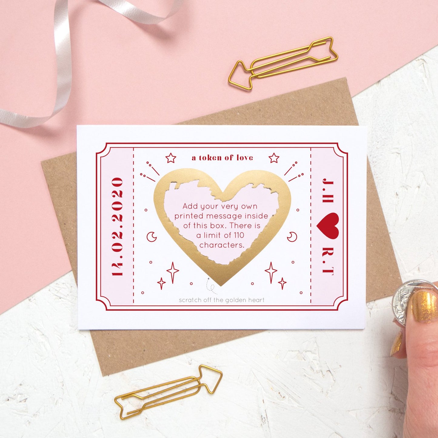 The personalised Love Token Scratch card with the printed message scratched off. Shot on a pink and white background with a hand in the corner.