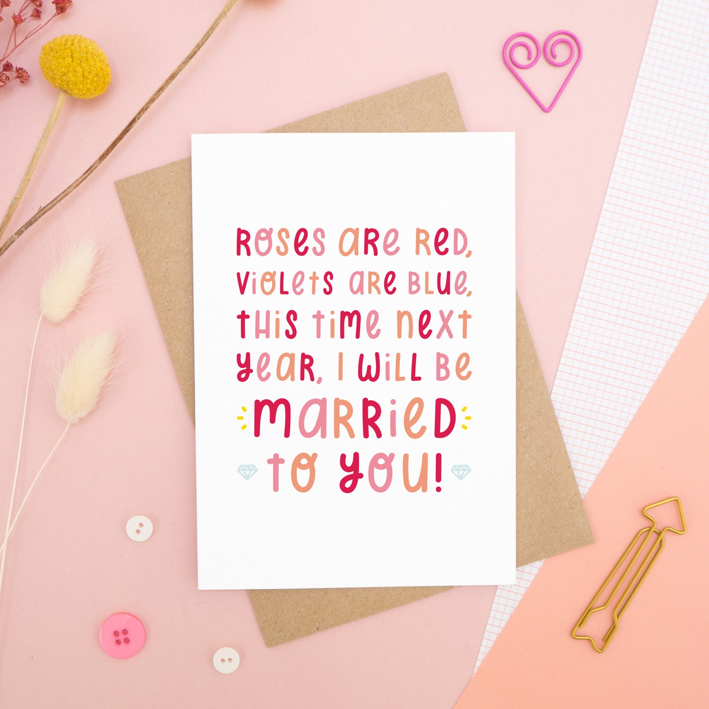 The 'this time next year' card photographed on a pink background with dried flowers, buttons and paper clips as props.