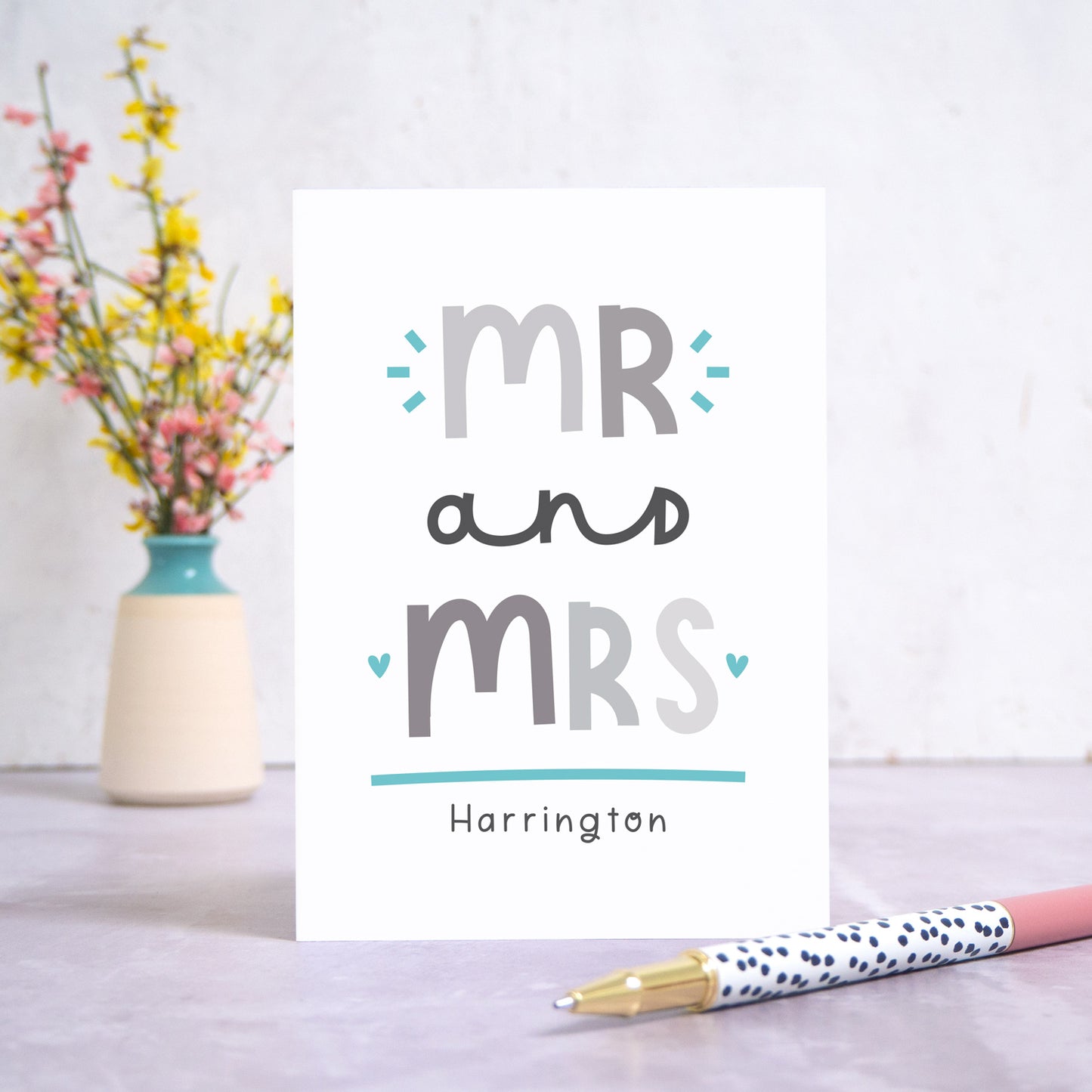 A Mr and Mrs personalised wedding congratulations card stood in front of a white background and a small vase of yellow and pink flowers. There is a white and black spotty pen in the foreground. This is the grey and blue version of the card.