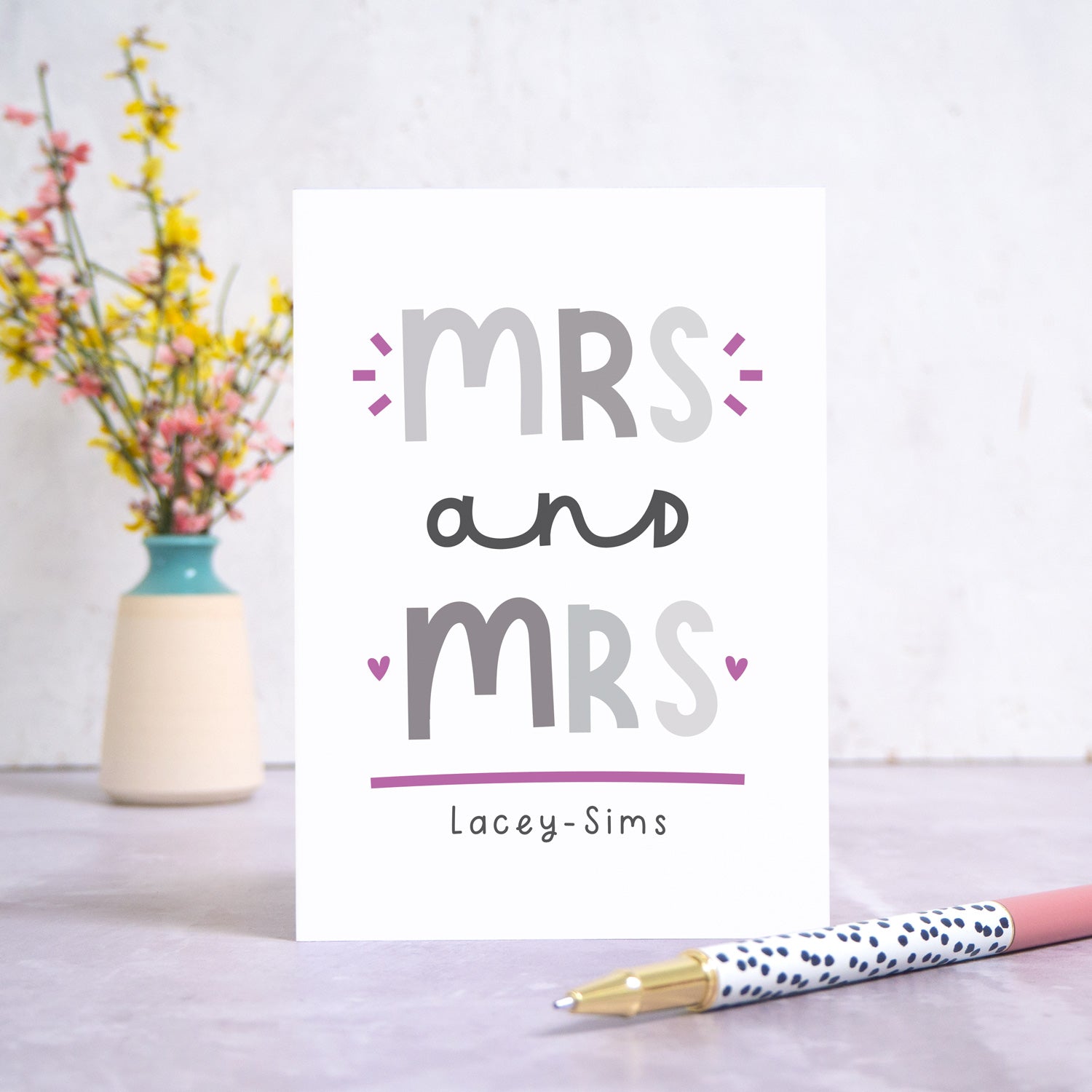 A Mrs and Mrs personalised wedding congratulations card stood in front of a white background and a small vase of yellow and pink flowers. There is a white and black spotty pen in the foreground. This is the grey and purple version of the card.