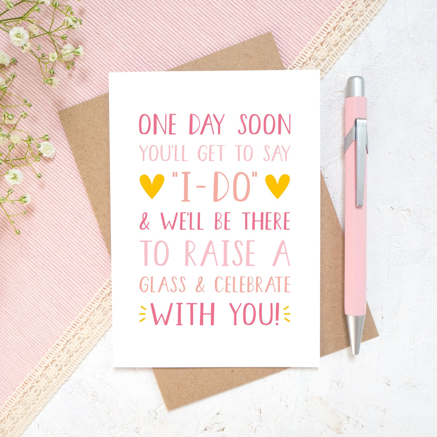 One day soon postponed wedding card in tones of pink. Photographed on a white and pink background with a hint of small flowers and a pen.