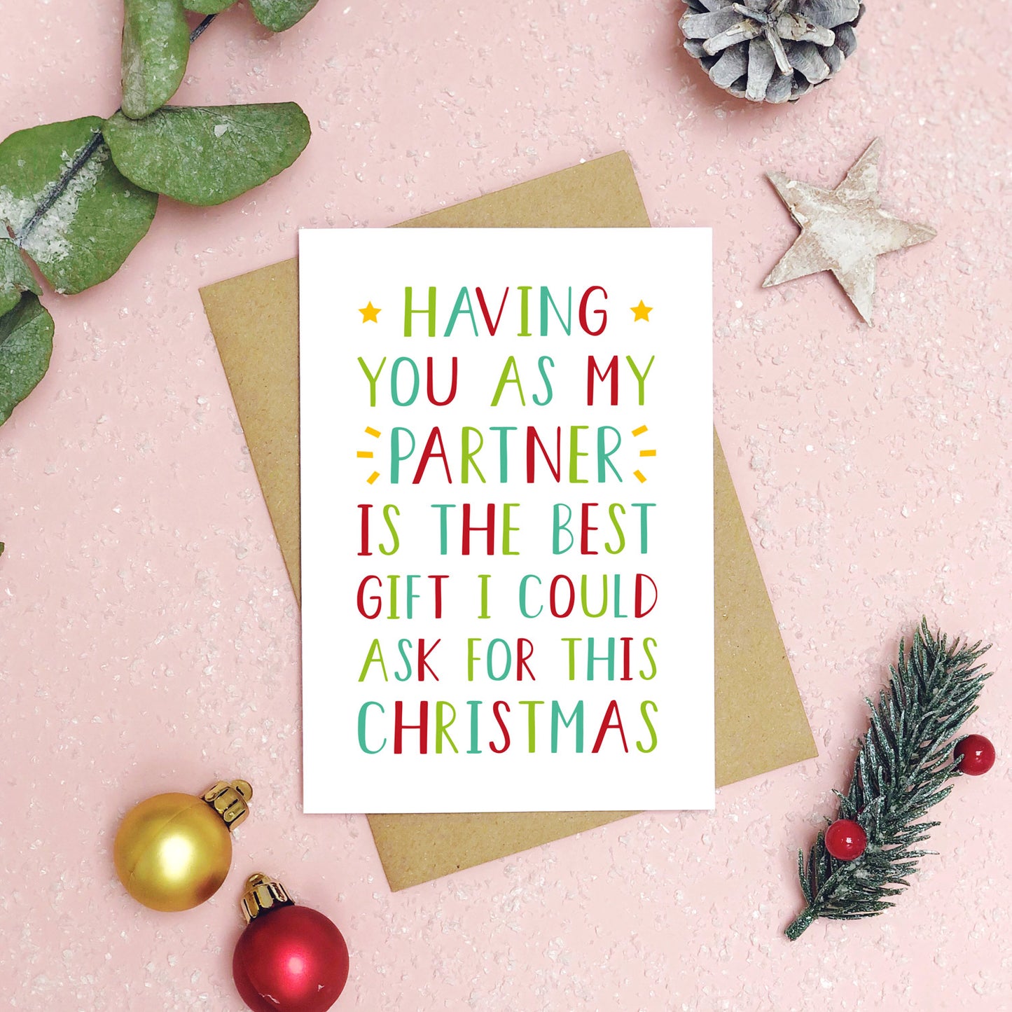 A best partner Christmas card as a flatlay on a pink background with foliage, baubles and festive decor. The card text is in green and red.