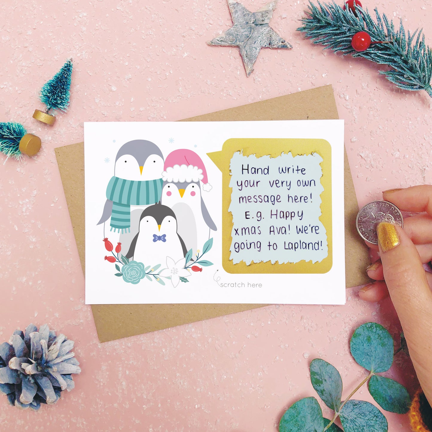 A personalised penguin family scratch card after the scratch panel has been scratched with a coin to reveal the hidden message.Shot on a pink background with grey and green festive props.