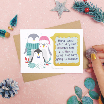 A personalised penguin family scratch card after the scratch panel has been scratched with a coin to reveal the hidden message.Shot on a pink background with grey and green festive props.