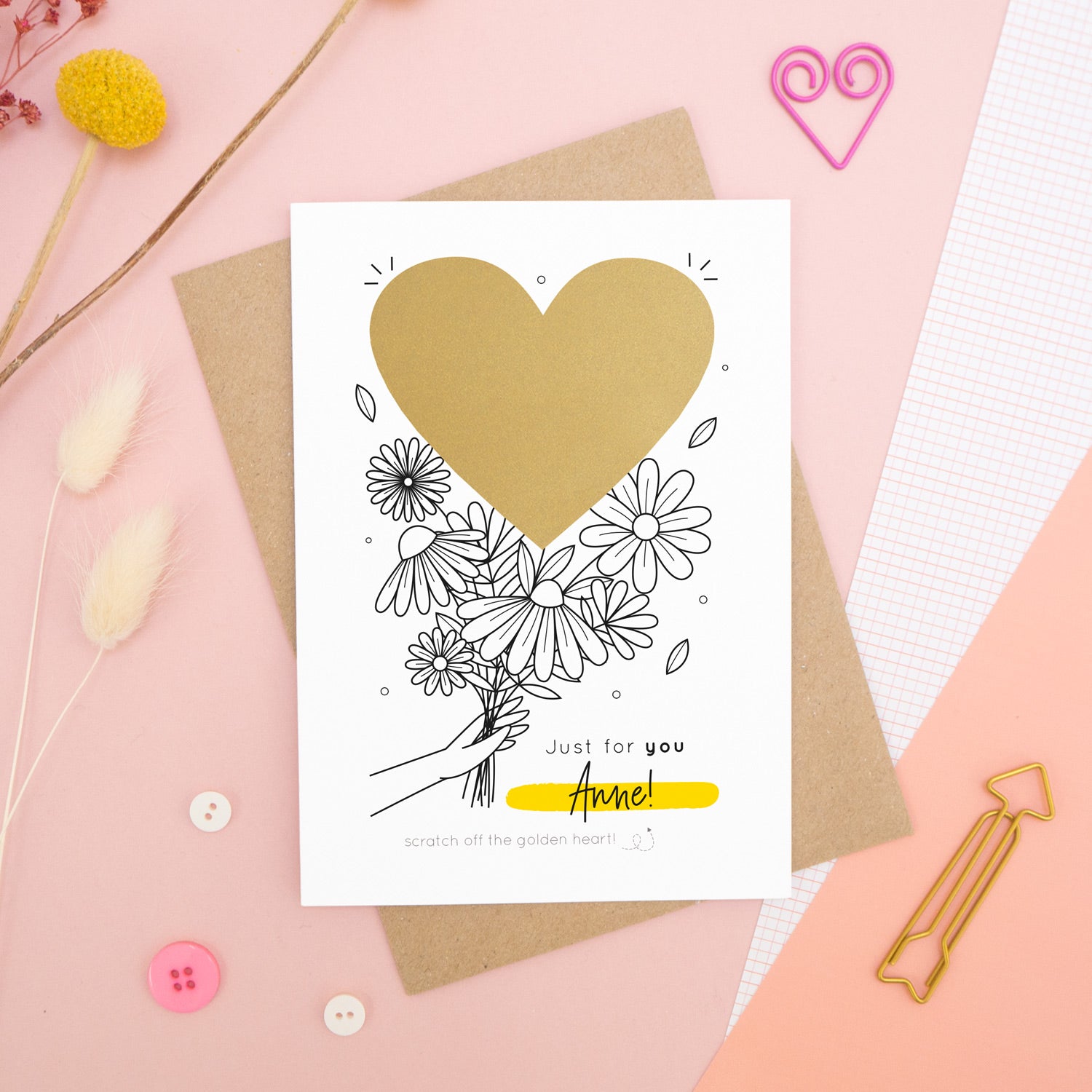 A personalised flower bouquet scratch card photographed on a pink background with floral props, paper clips, and buttons. This card shows a black and white floral illustration and the golden heart before it is scratched off to reveal the secret message!
