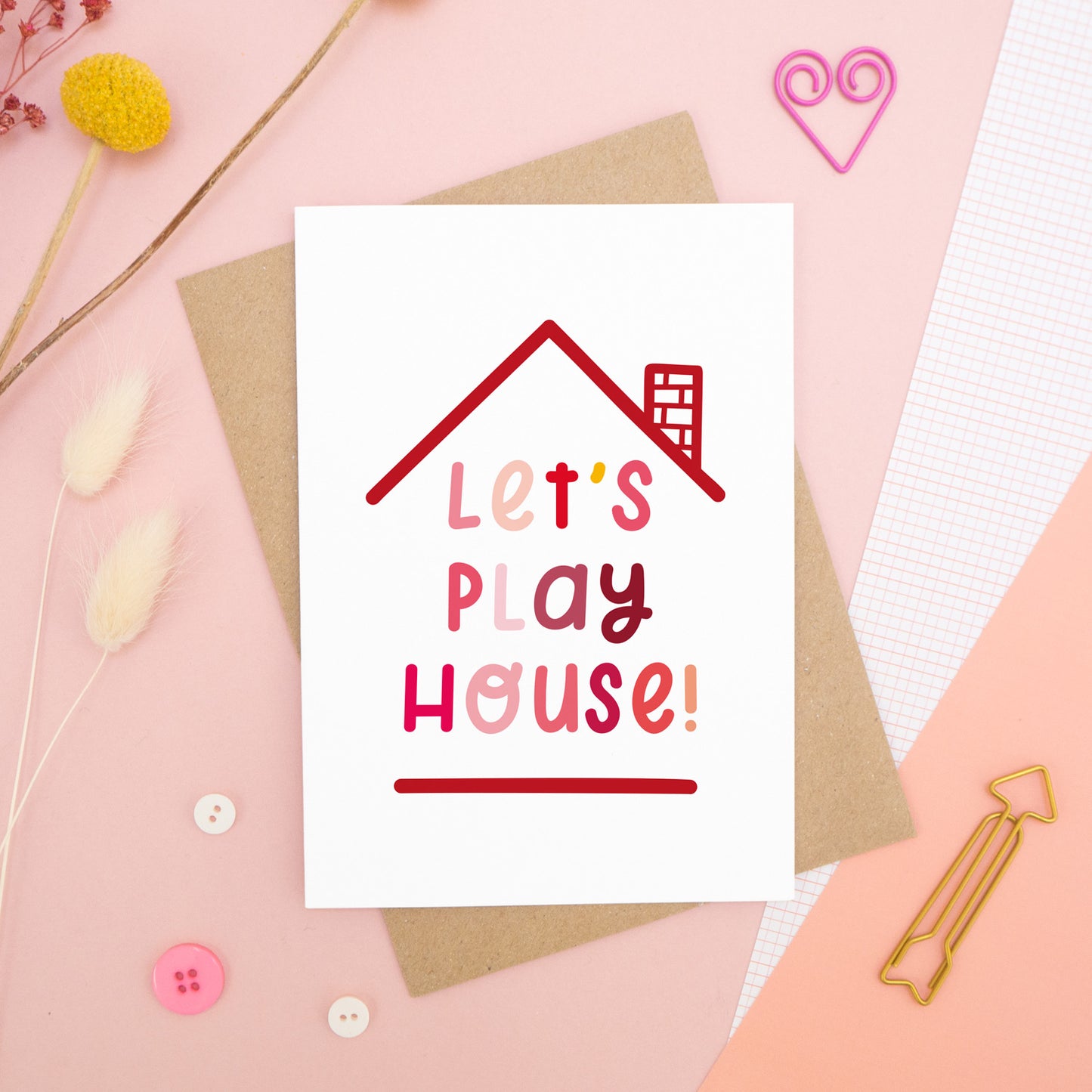The 'let's play house' card photographed on a pink background with dried flowers, buttons and paper clips as props.