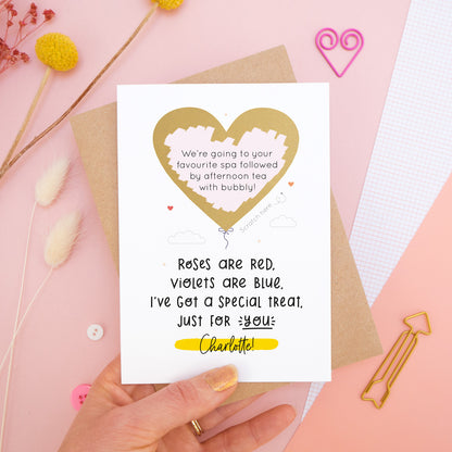 A personalised anniversary scratch card photographed on a pink background with floral props, paper clips, and buttons. This card shows the golden heart after it has been scratched off to reveal the secret message!