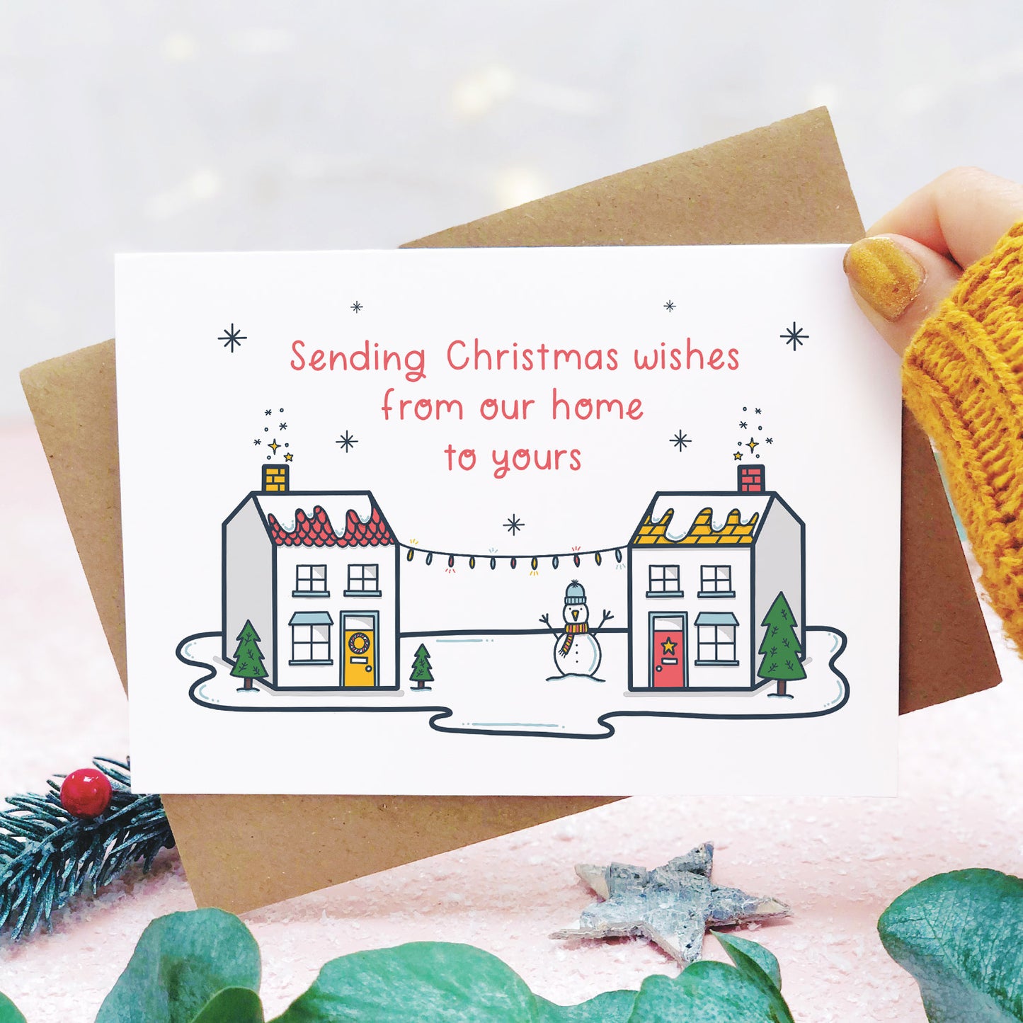 A sending wishes from our home to yours card photographed on a pink background, and being held on the right hand side with a yellow sleeve in view, with grey and green foliage. The card features two little houses connected by fairy lights.