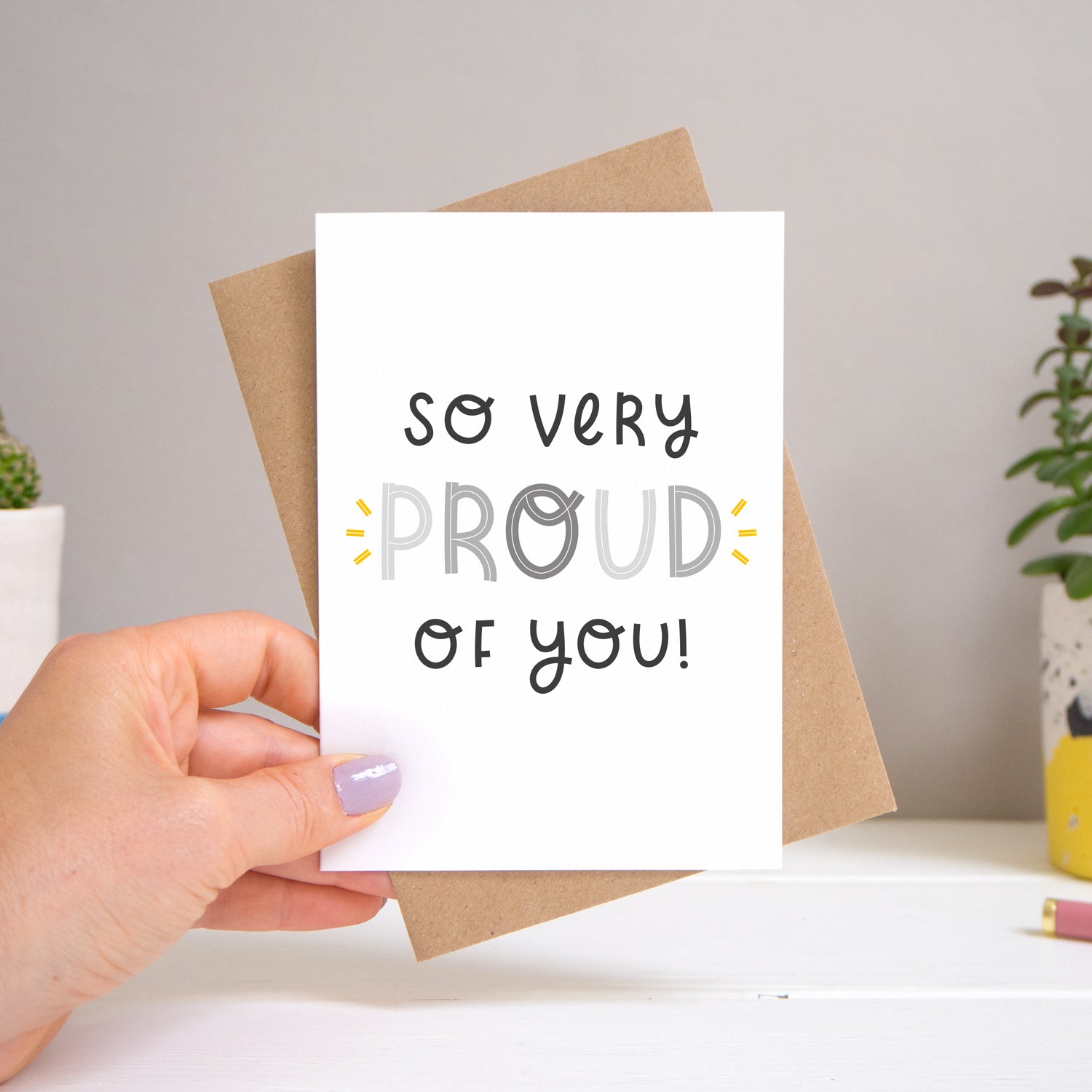 A ‘so very proud of you’ card held over a warm grey and white background with potted plants peeping the sides. Behind the card is a kraft brown envelope that comes with the card. The text on this version of the card is in varying tones of grey.