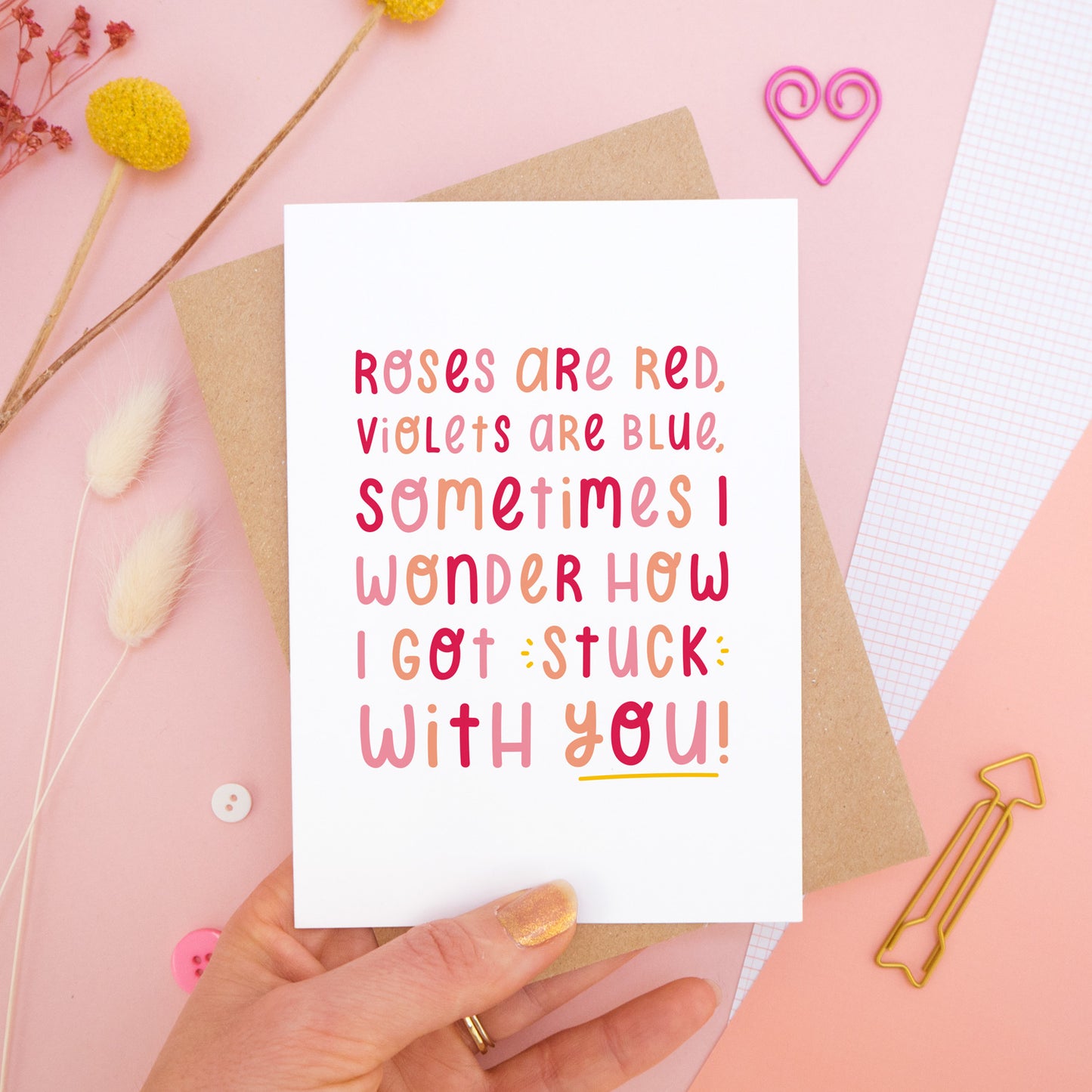 The 'stuck with you' card photographed on a pink background with dried flowers, buttons and paper clips as props. The card itself is being held above the scene.