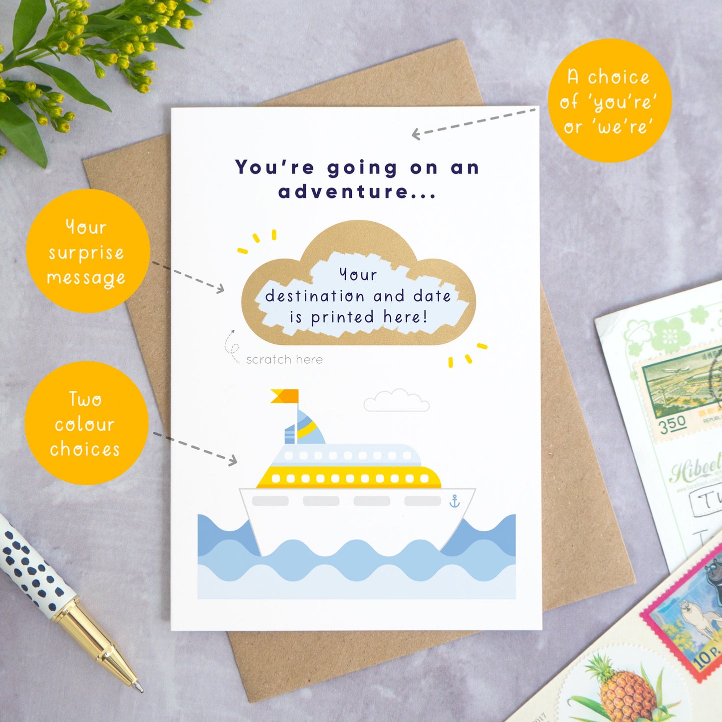 A personalised cruise ship holiday announcement scratch card photographed on a grey concrete background with greenery, a pen and postcards around the edge. The card is set upon a kraft brown envelope. The card is in the blue colour palette and the cloud has been scratched off to reveal the custom travel plans. The orange circles demonstrate what can be changed on the card. For example the wording, colour and custom scratch message.