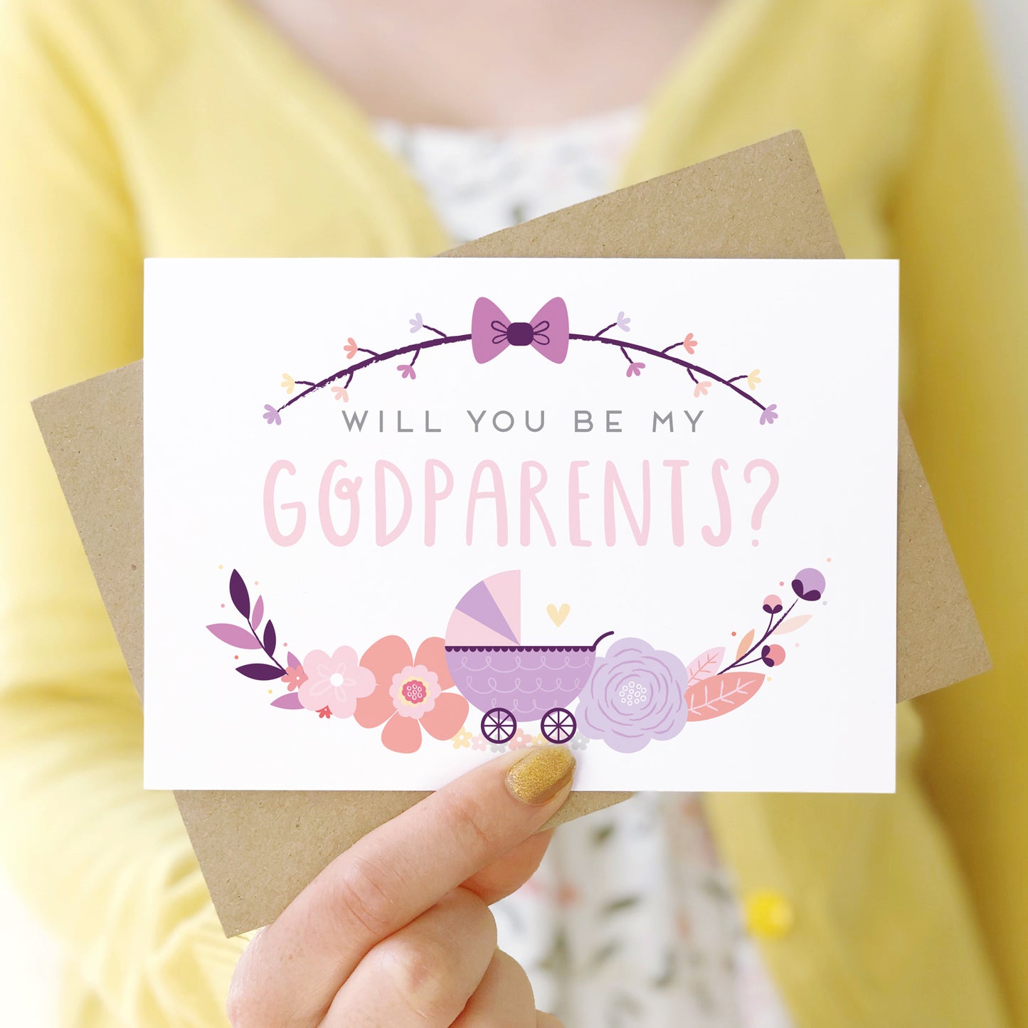 A will you be my godparents card being held in front of a white dress and yellow cardigan. The design features a pram, simple florals and the all important question. This is the purple palette.