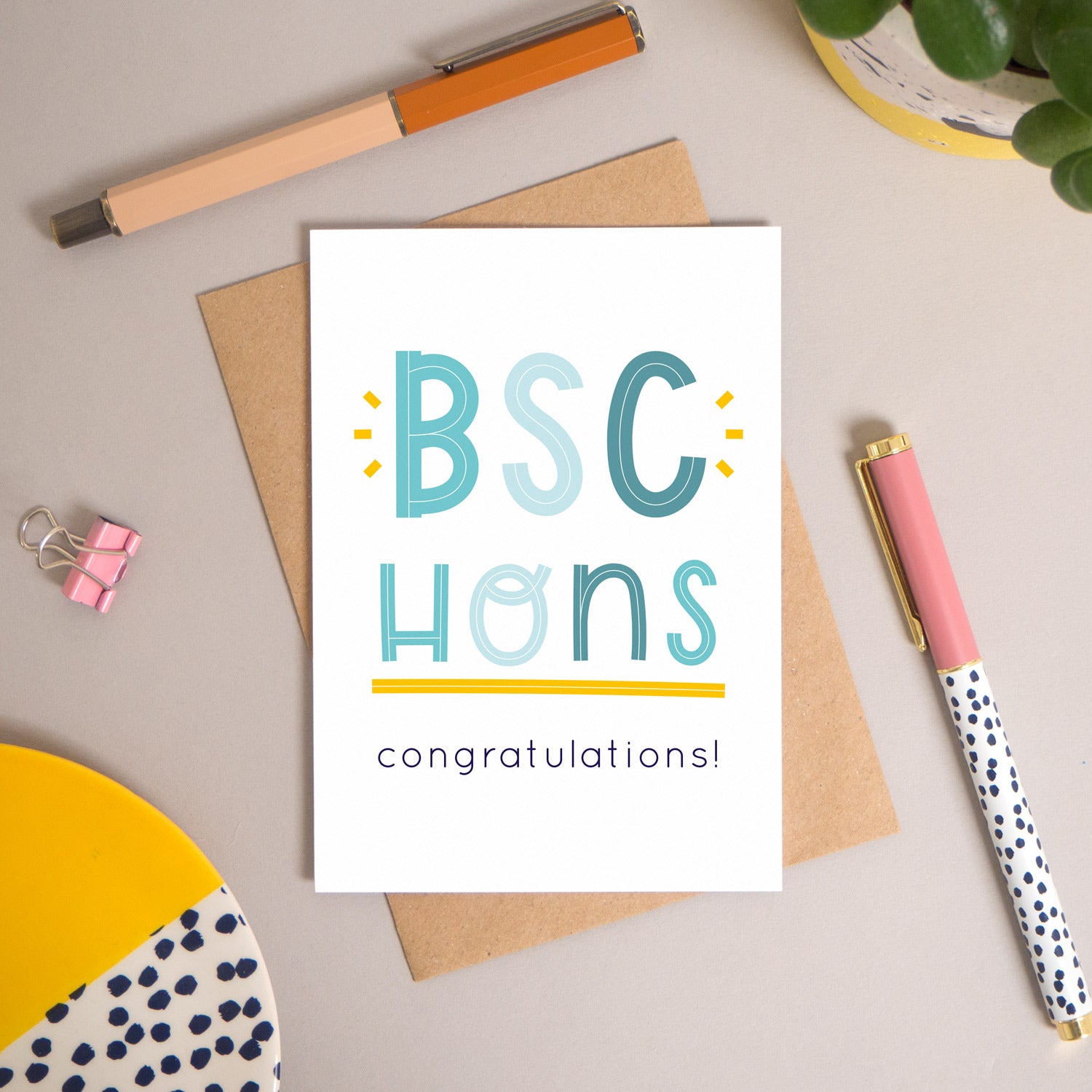 This BSc Hons graduation congratulations card has been shot flatlay style looking directly over the top of the card. It is lying flat on it’s kraft brown envelope, on a warm grey background. Surrounding the card are pens, a clip, a plant and a trinket dish. This version of the card is in varying tones of navy and blue.
