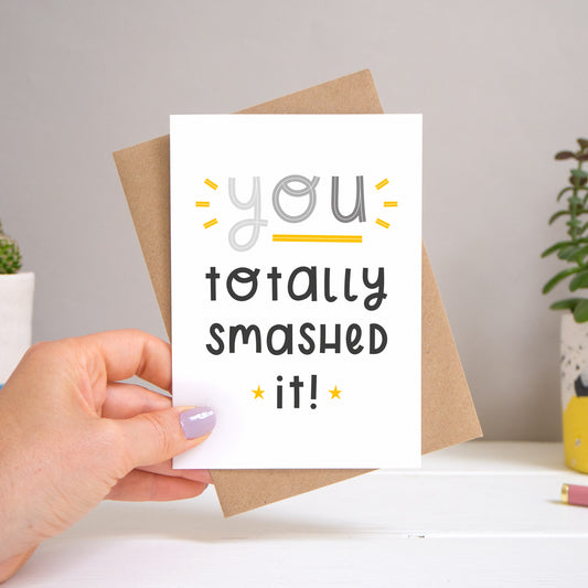 A ‘you totally smashed it’ card held over a warm grey and white background with potted plants peeping the sides. Behind the card is a kraft brown envelope that comes with the card. The text on this version of the card is in varying tones of grey.