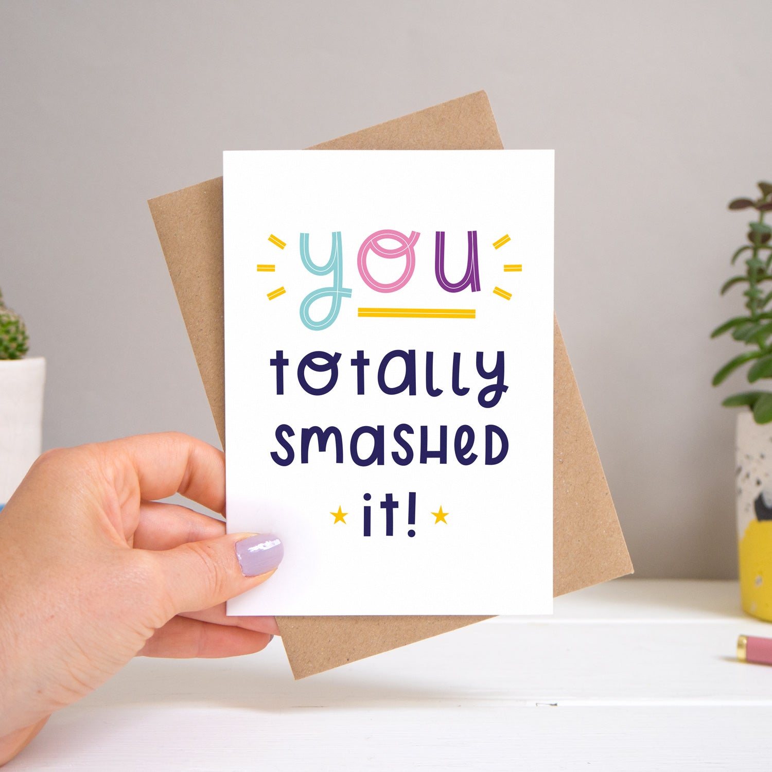 A ‘you totally smashed it’ card held over a warm grey and white background with potted plants peeping the sides. Behind the card is a kraft brown envelope that comes with the card. The text on this version of the card is in varying tones of navy and pink, purple and blue.