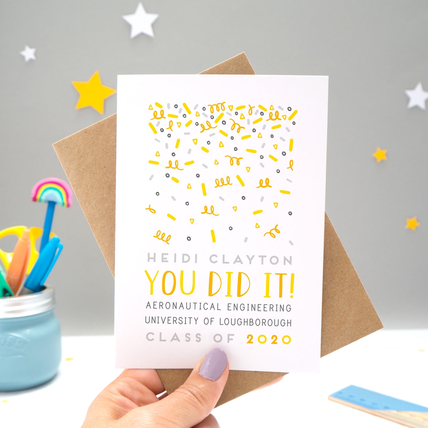 A personalised graduation card designed and made by Joanne Hawker in her somerset studio being held against a kraft brown envelope over a grey background with yellow and white stars. The confetti illustration and text is in varying tones of grey, yellow and orange.