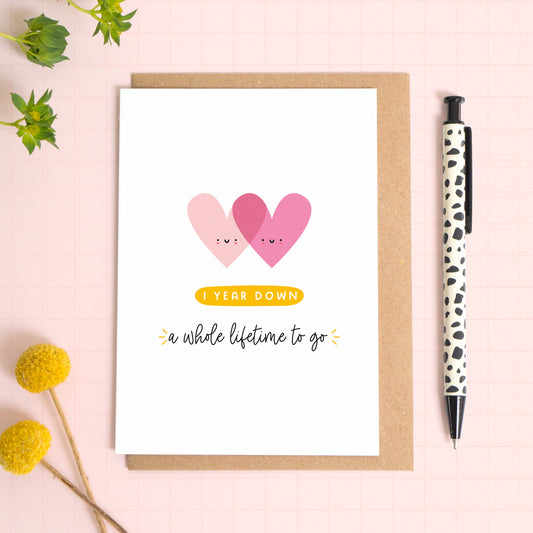 A ‘1 year down - a whole lifetime to go’ card for a 1st wedding anniversary which has been photographed on a Kraft brown envelope upon a pink background surrounded by florals and a pen for scale.