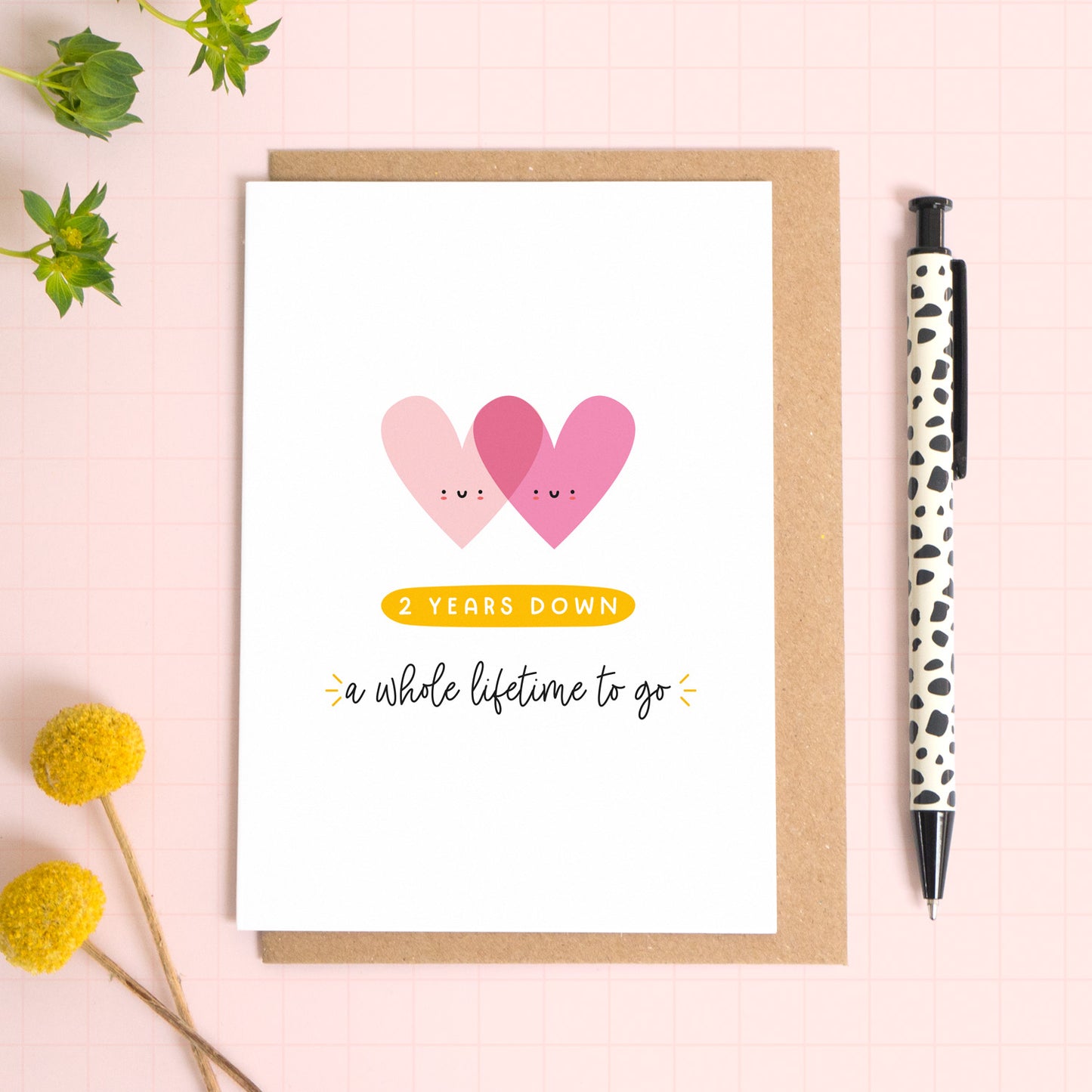 A ‘2 years down - a whole lifetime to go’ card for a 2nd wedding anniversary which has been photographed on a Kraft brown envelope upon a pink background surrounded by florals and a pen for scale.