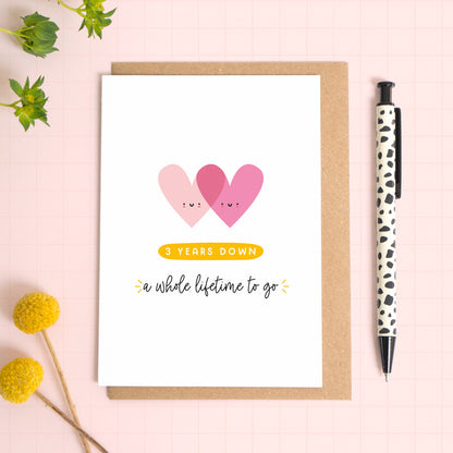 A ‘3 years down - a whole lifetime to go’ card for a 3rd wedding anniversary which has been photographed on a Kraft brown envelope upon a pink background surrounded by florals and a pen for scale.