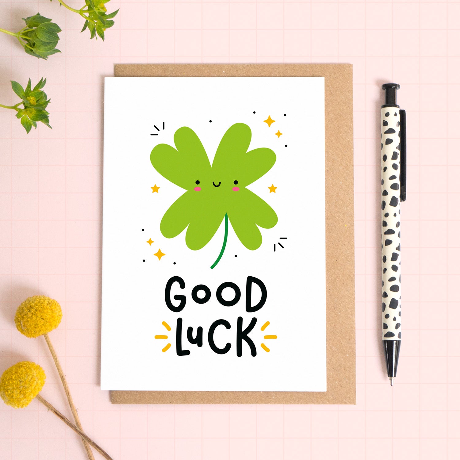 A good luck card with an illustration of a four leaf clover, and the phrase ‘good luck’ printed on the front of the card. The card has been photographed on it's kraft brown envelope on a pink surface with foliage and a pen for scale.