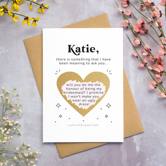 The personalised question scratch card has been photographed lying on top of a Kraft brown envelope which is on top of a grey surface surrounded by colourful flowers. The gold heart scratch panel has been scratched away to reveal the surprise question.
