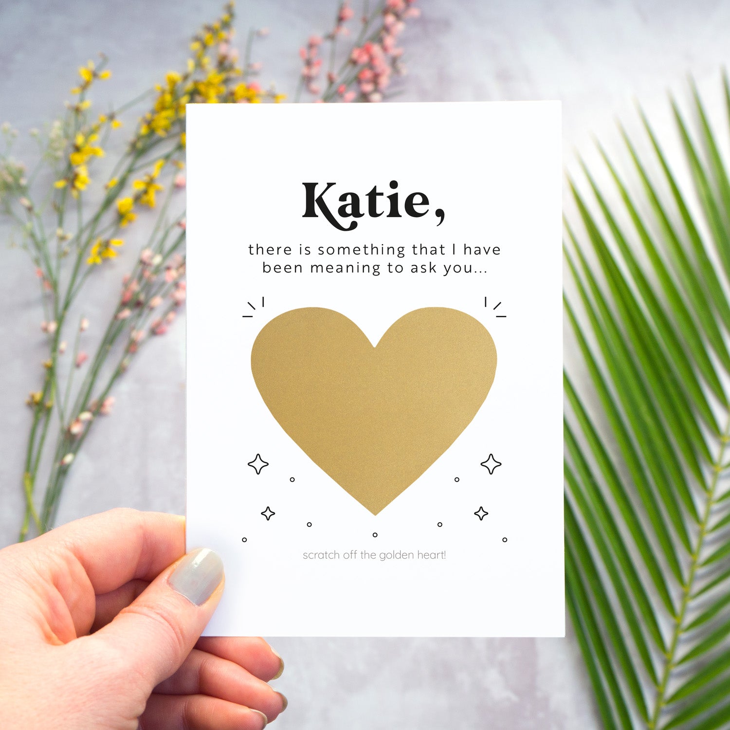 This image shows how the personalised question scratch card looks before the recipient scratches away the heart. In this image the card is being held over a grey surface which has a variety of colour flowers and foliage in the background. The heart has not yet been scratched away.