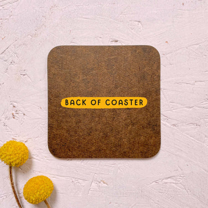 The back of all of the coasters come as 'raw hardboard'. This is a smooth surface. This coaster has been photographed on a pink surface with two yellow flowers in the bottom left corner.