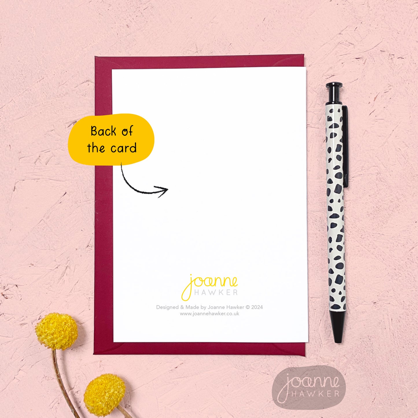 The back of the greetings card showing the Joanne Hawker logo, copyright info and website. The card has a red envelope and it has been photographed on a pink background next to yellow flowers and a pen for scale.