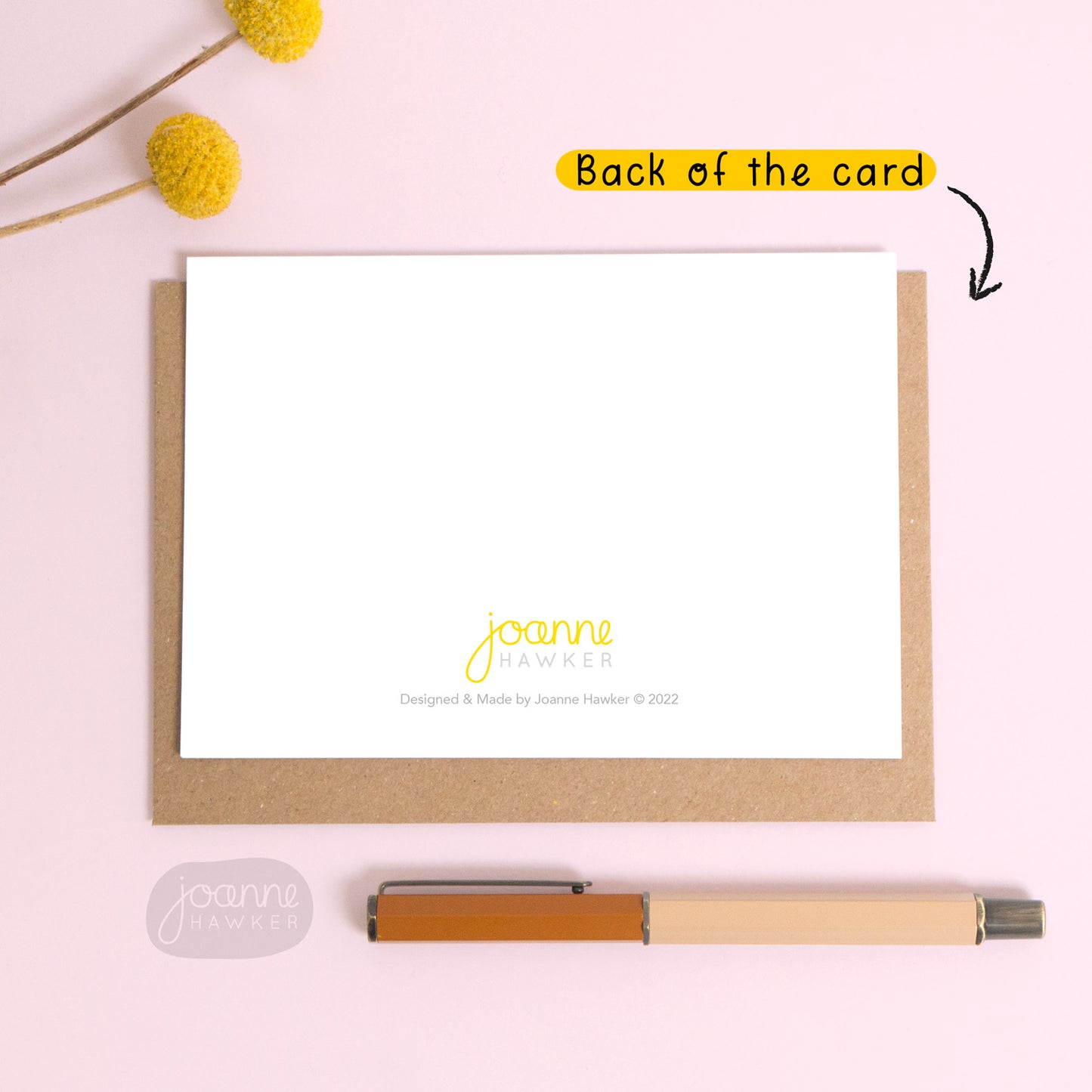 This image demonstrates the back of the greetings card. The card is in landscape format and shows the ‘Joanne Hawker’ logo and copyright information bottom centre. The card is laid down on top of a Kraft brown envelope and photographed on a pink background with text pointing to the card reading “back of the card”.