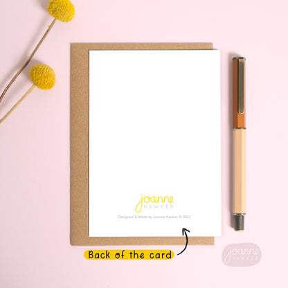 This image demonstrates the back of the greetings card. The card is in portrait format and shows the ‘Joanne Hawker’ logo and copyright information bottom centre. The card is laid down on top of a Kraft brown envelope and photographed on a pink background with text pointing to the card reading “back of the card”.