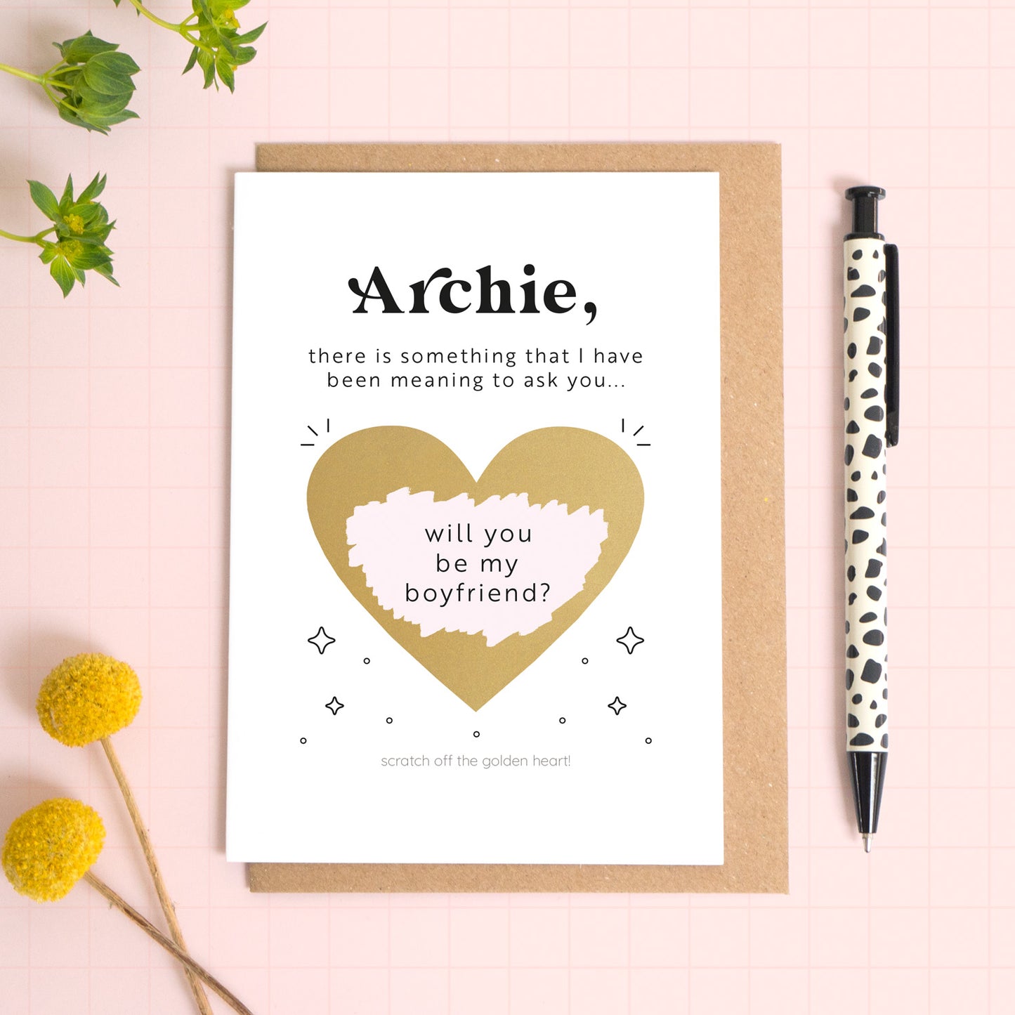 A ‘will you be my boyfriend’ scratch and reveal card photographed lying on a Kraft brown envelope, set upon a pink background surrounded by foliage and a pen for scale. The gold heart has been scratched off to reveal the personalised question.