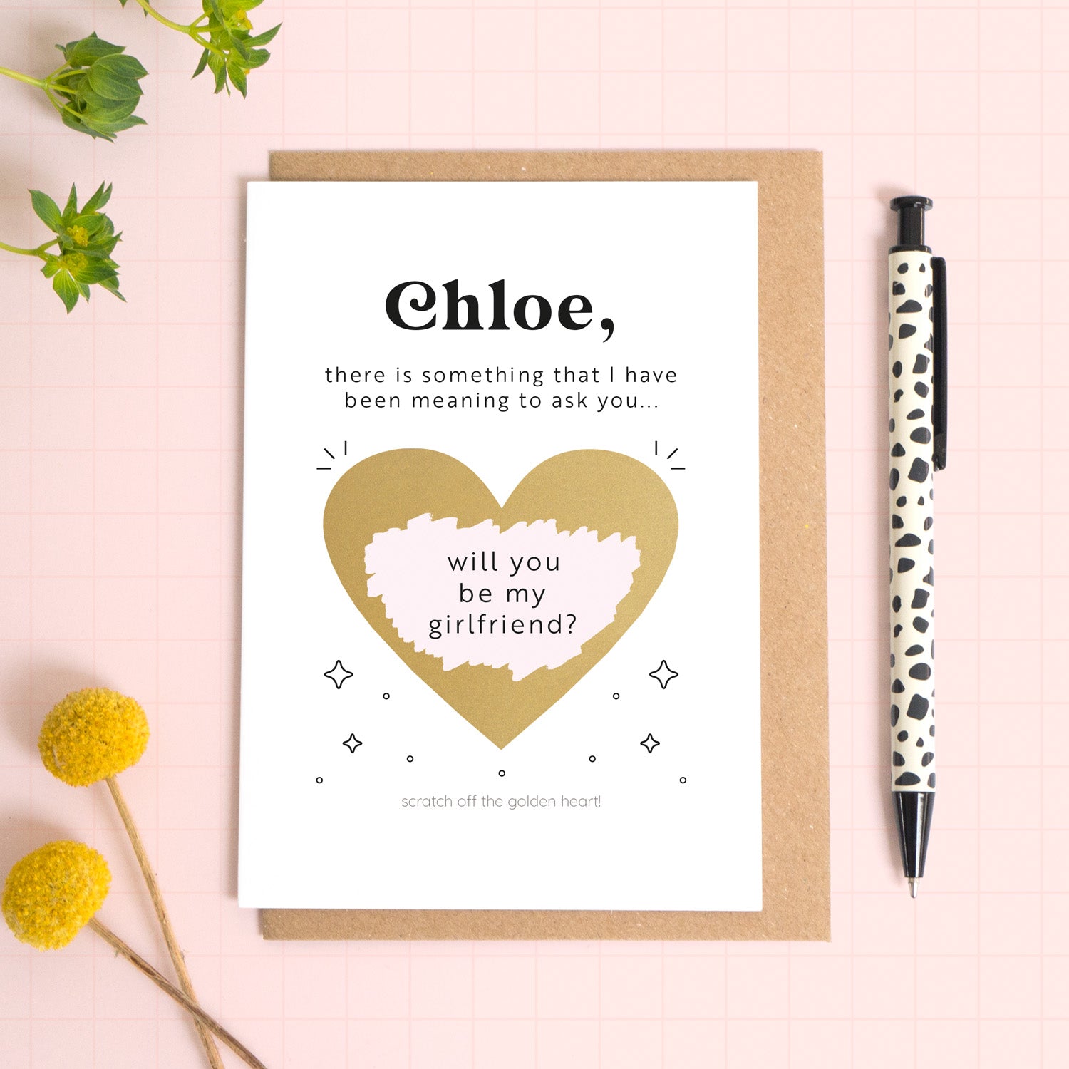 A ‘will you be my girlfriend’ scratch and reveal card photographed lying on a Kraft brown envelope, set upon a pink background surrounded by foliage and a pen for scale. The gold heart has been scratched off to reveal the personalised question.