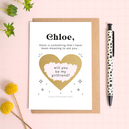 A ‘will you be my girlfriend’ scratch and reveal card photographed lying on a Kraft brown envelope, set upon a pink background surrounded by foliage and a pen for scale. The gold heart has been scratched off to reveal the personalised question.