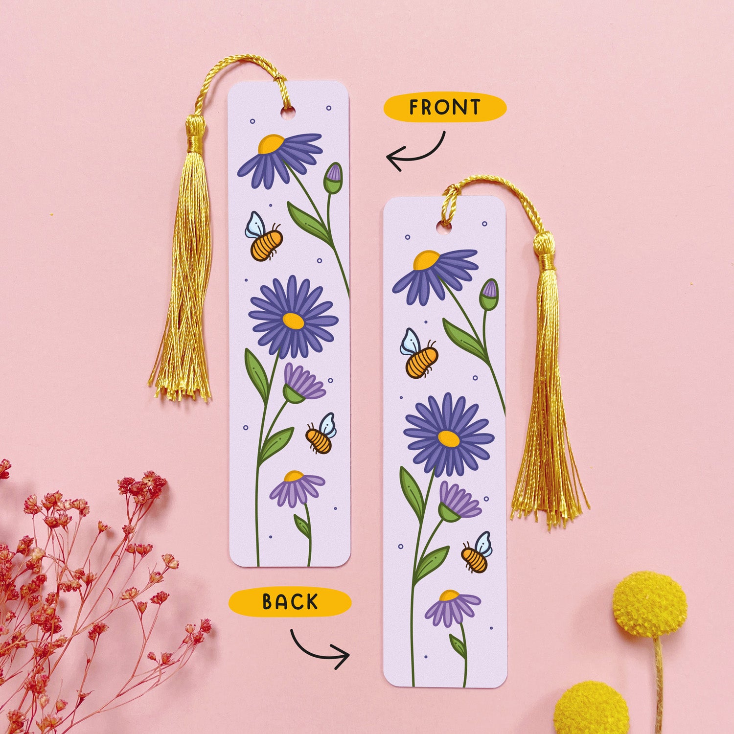 An image depicting the front and the back of the double sided metal bookmark which has been photographed on a pink background with dried flowers. Each bookmark has a gold tassel. The front and the back of the bookmark are printed with the same design of purple aster flowers and bumble bees.