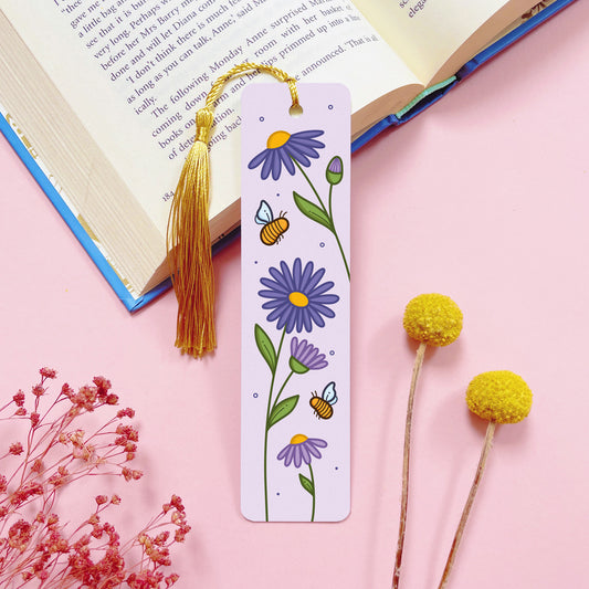 An aster flowers and bees themed, double sided, aluminium bookmark with a gold tassel which has been photographed on a pink background with an open book and dried flowers.